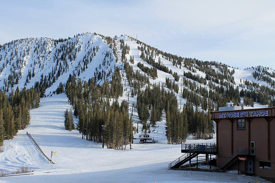 The Mt Rose front side in full sun - cut runs fanning down a forested peak, a chairlift crossing the frame, and the base lodge with its 'MT ROSE SKI TAHOE' sign at right.