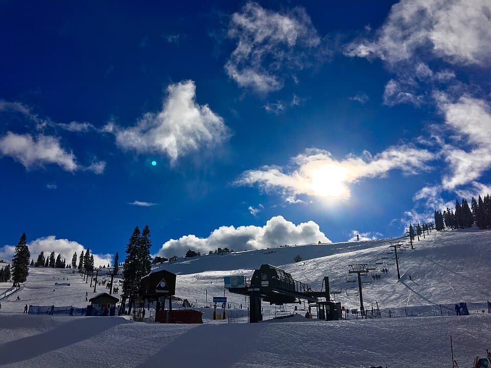Tahoe Donner's downhill slope on a bluebird day, chairlift and lift shack in silhouette against fresh snow with the sun flaring over the ridge.